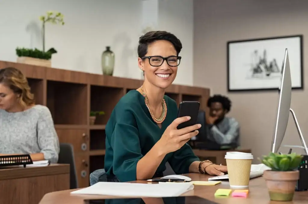 Smiling businesswoman using smartphone at desk in modern office workspace with computer and coffee cup.