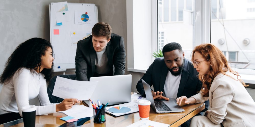 Business briefing meeting. Successful group of people looking at financial graphs and diagrams, brainstorming together. Multiethnic male and female employee discuss ideas, working on new project