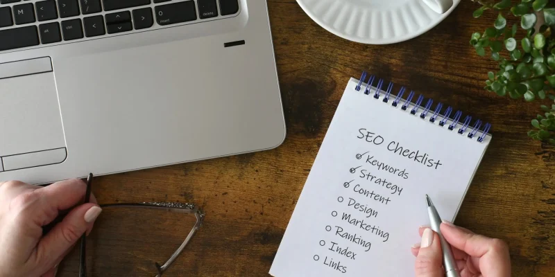 Person reviewing SEO checklist with keywords, content strategy, ranking, and links on a notepad next to a laptop on a wooden desk.