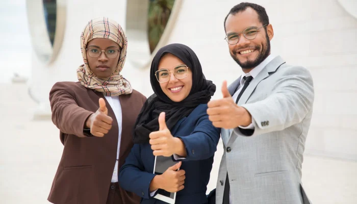 Multicultural business group posing and making like gesture. Businessman and Muslim businesswomen standing outdoors, showing thumb up and smiling at camera. Successful business team concept