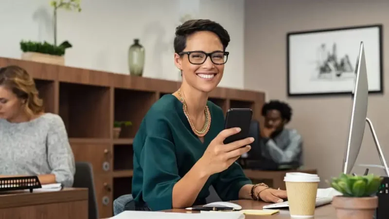 Smiling businesswoman using smartphone at desk in modern office workspace with computer and coffee cup.